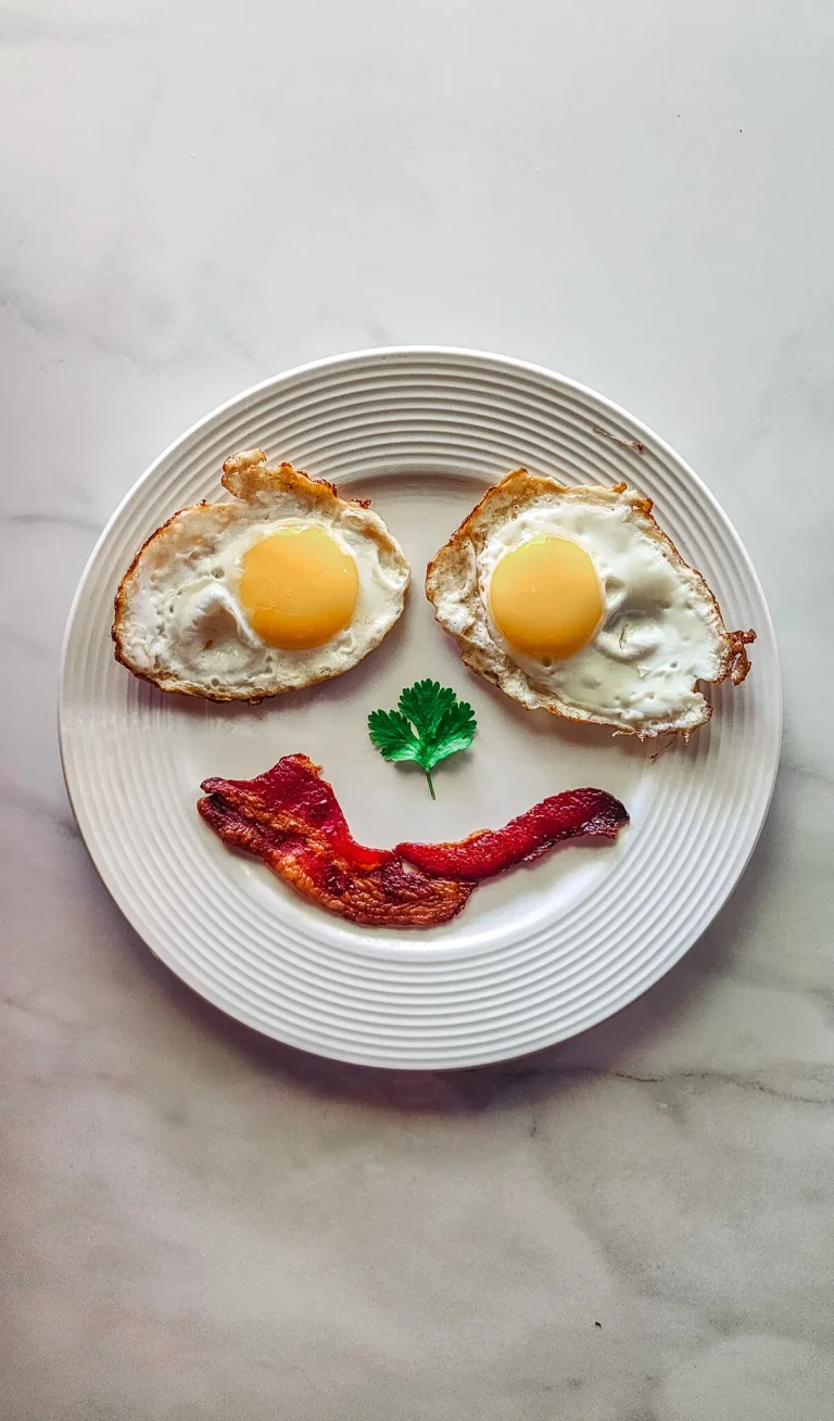 A breakfast plate with sunny-side-up eggs, bacon, and a sprig of parsley arranged as a smiley face, served alongside a stack of pancakes and crispy tater tots. Title: A Goofy Movie-Inspired Breakfast – Max’s Bacon & Eggs