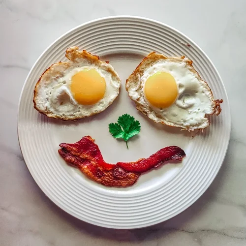 A breakfast plate with sunny-side-up eggs, bacon, and a sprig of parsley arranged as a smiley face, served alongside a stack of pancakes and crispy tater tots. Title: A Goofy Movie-Inspired Breakfast – Max’s Bacon & Eggs