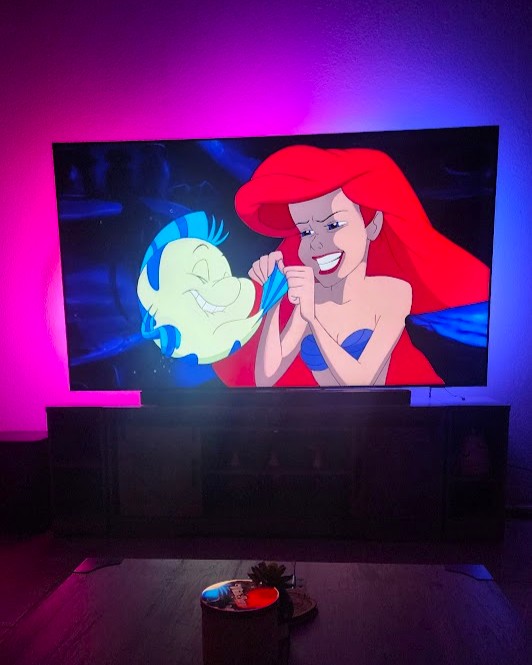 A scene from The Little Mermaid featuring Ariel and Flounder on a Samsung TV with Philips Hue lights creating a vibrant pink and purple glow.