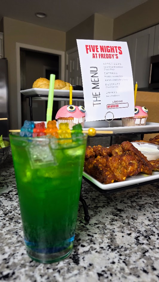 Neon green-blue Freddy Fizz cocktail topped with rainbow gummy bears, next to the Five Nights at Freddy’s menu card.