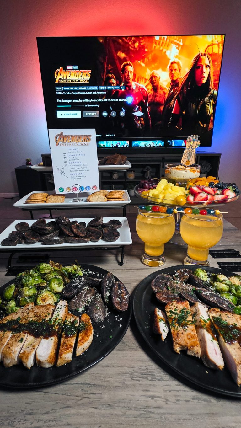 Avengers Infinity War title screen on TV with full dinner spread including pork chop platters, cocktails, and Infinity Stones Board on coffee table