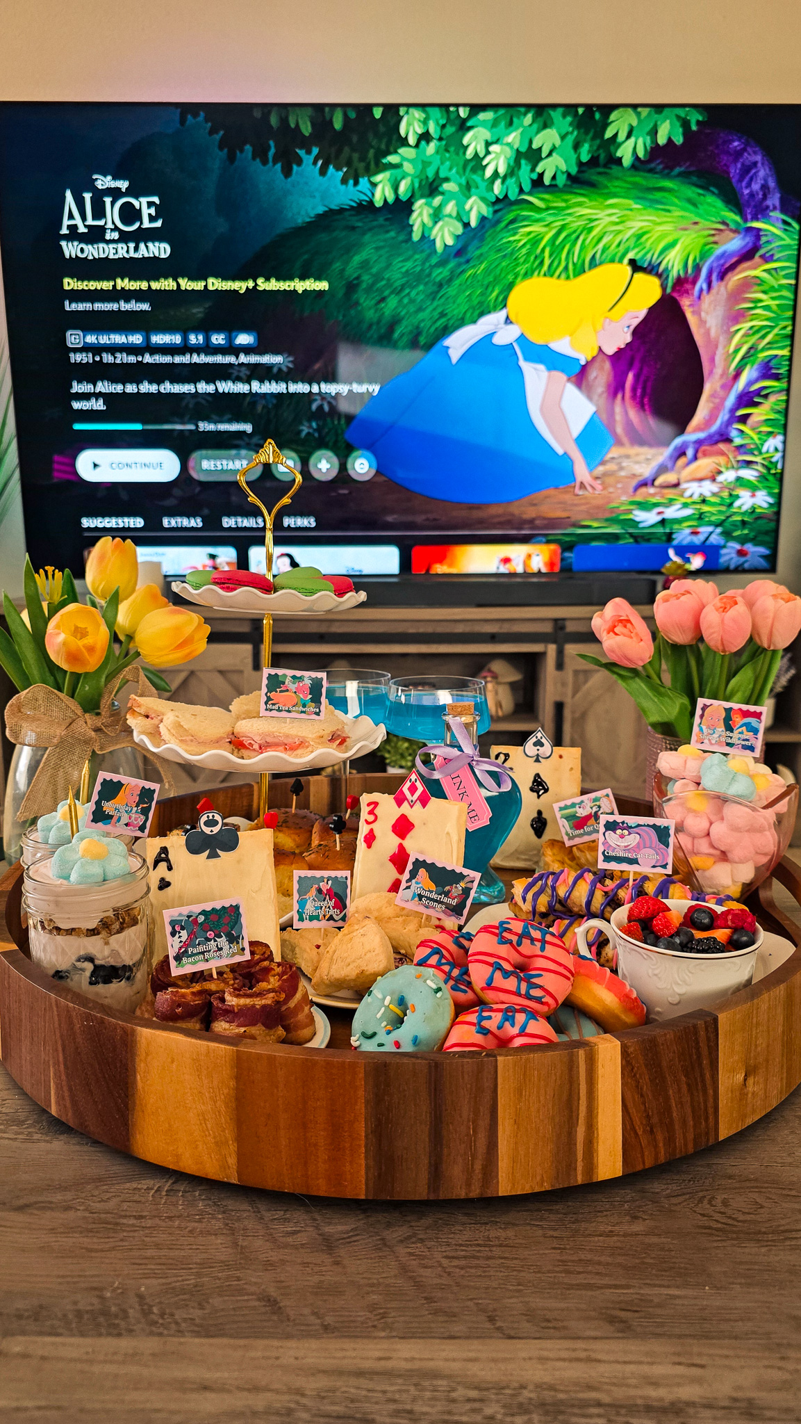 Alice in Wonderland tea party brunch board on a lazy Susan with a tiered tray in front of a TV showing the Alice in Wonderland title card